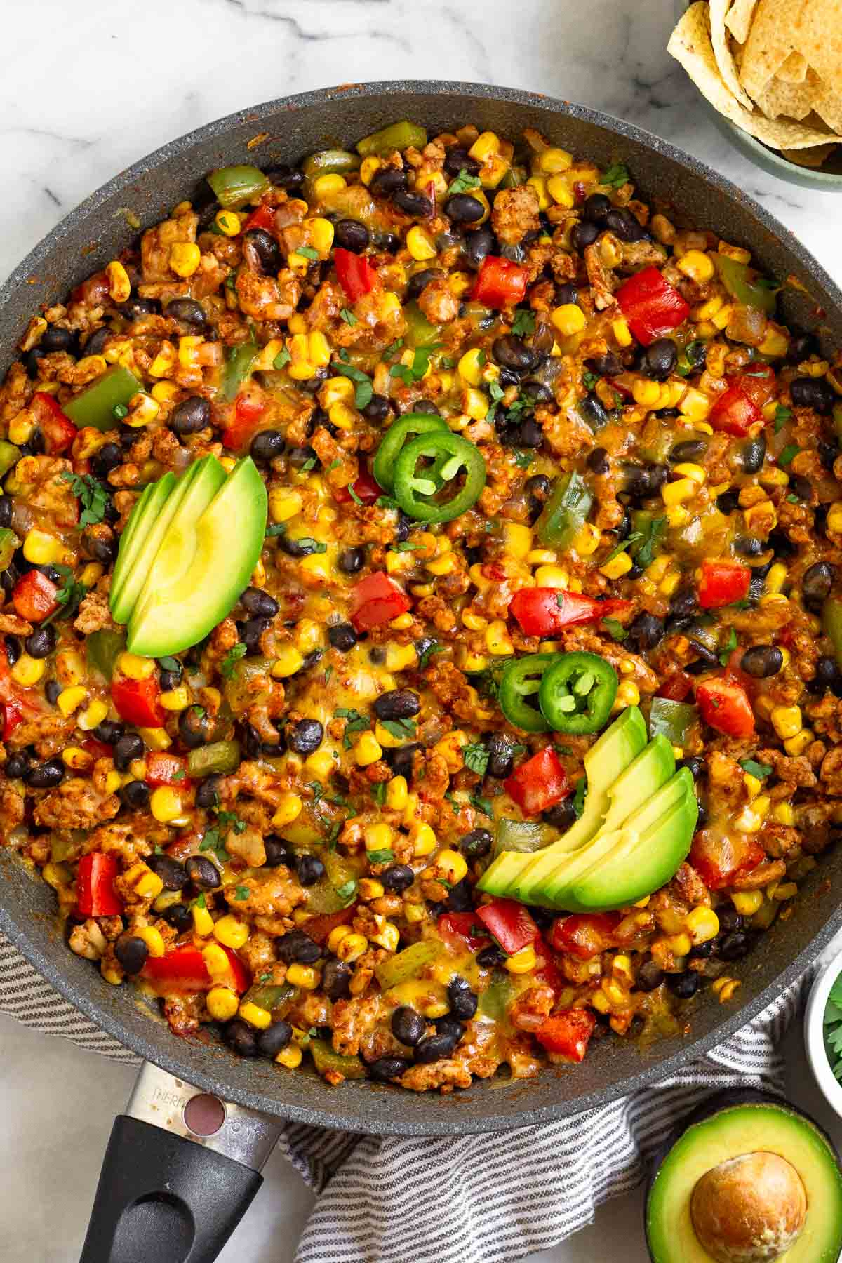 Chicken and corn skillet with veggies and black beans topped with melted cheese. It is garnished with sliced avocado, sliced jalapeños, and cilantro. Next to the skillet is half an avocado and a bowl of tortilla chips.