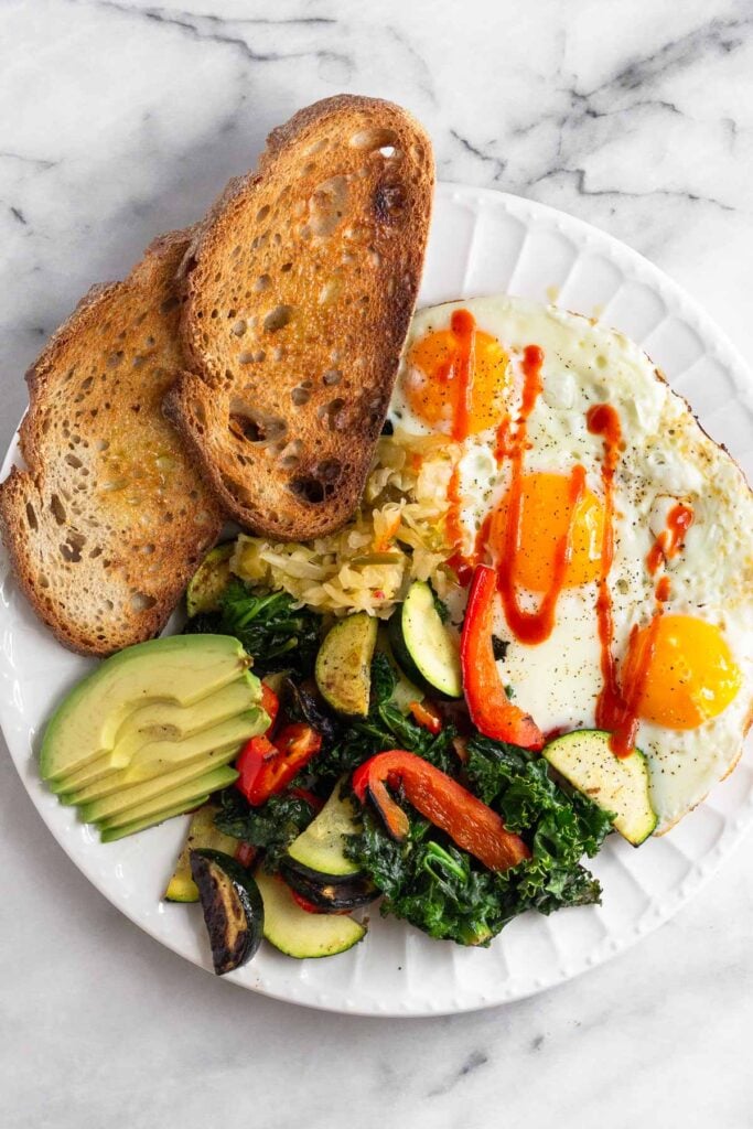 High protein breakfast with 3 fried eggs, sautéed veggies, sliced avocado, and 2 pieces of toast on a white plate.