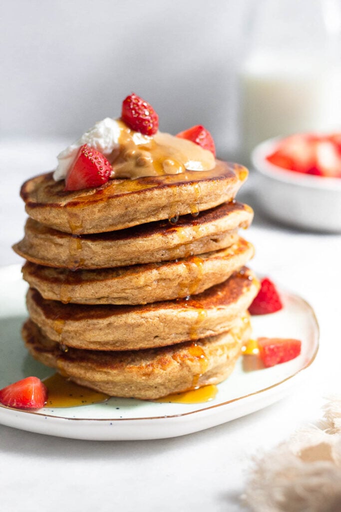 Protein pancakes stacked on a plate topped with yogurt, peanut butter, strawberries, and maple syrup. Behind them is a bowl of strawberries and a jar of milk.