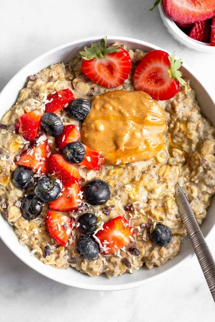 Bowl of cauliflower oatmeal topped with berries and peanut butter with a spoon coming out of it. There is a small bowl of cut up strawberries next to it.