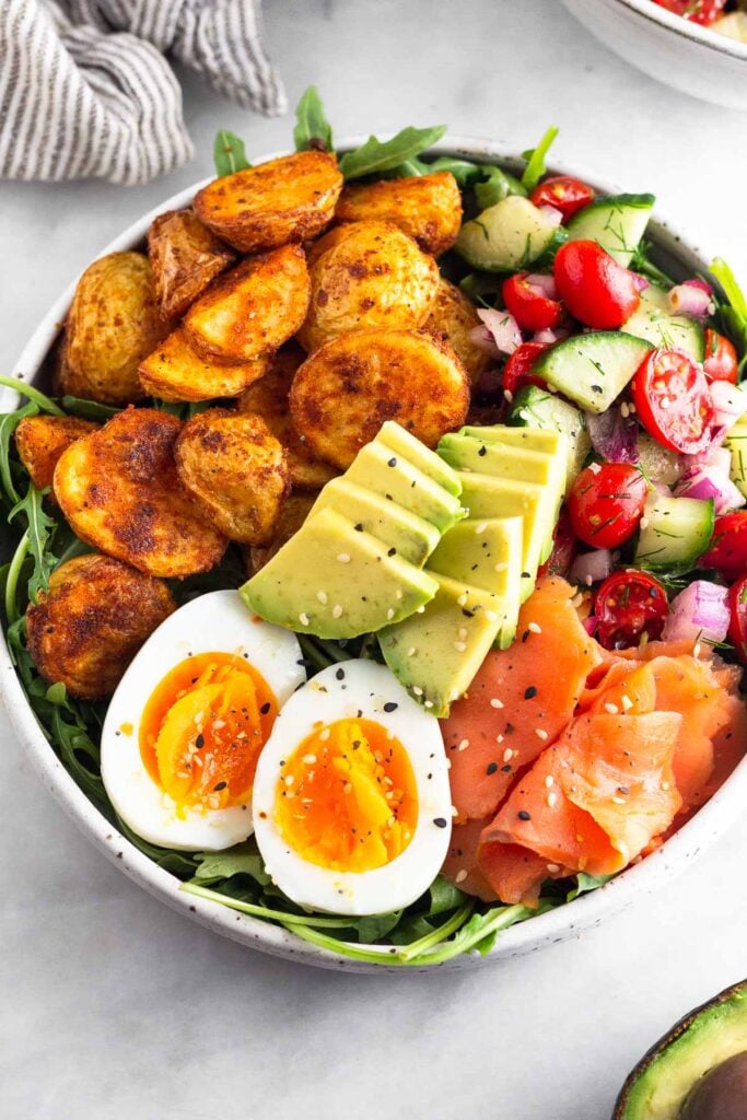 Protein breakfast bowl recipe with greens, roasted potatoes, tomato and cucumber salad, smoked salmon, hard boiled egg, and avocado topped with everything but the bagel seasoning.