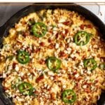 Cast iron skillet filled with jalapeño popper chicken casserole topped with melted cheese, crumbled pretzels, and sliced jalapeños.