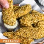 A plate of pistachio crusted chicken tenders with a small dish of ranch. One of the chicken tenders is being dipped in the ranch. Off in the background is bowl of shelled pistachios.