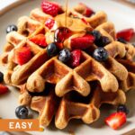 Protein waffles on a plate topped with berries and nut butter. Maple syrup is doing poured on top of them.