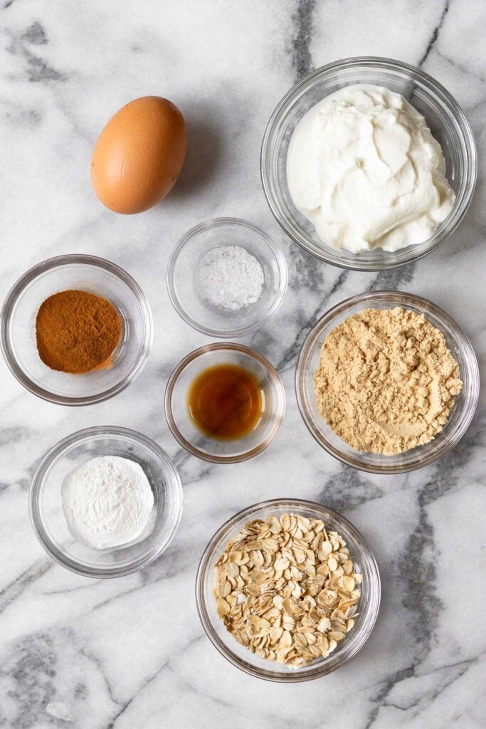 White marble counter with a bowl of greek yogurt, a bowl of protein powder, a bowl of rolled oats, a bowl baking powder, a bowl of vanilla extract, a bowl of salt, a bowl of cinnamon, and one brown egg.