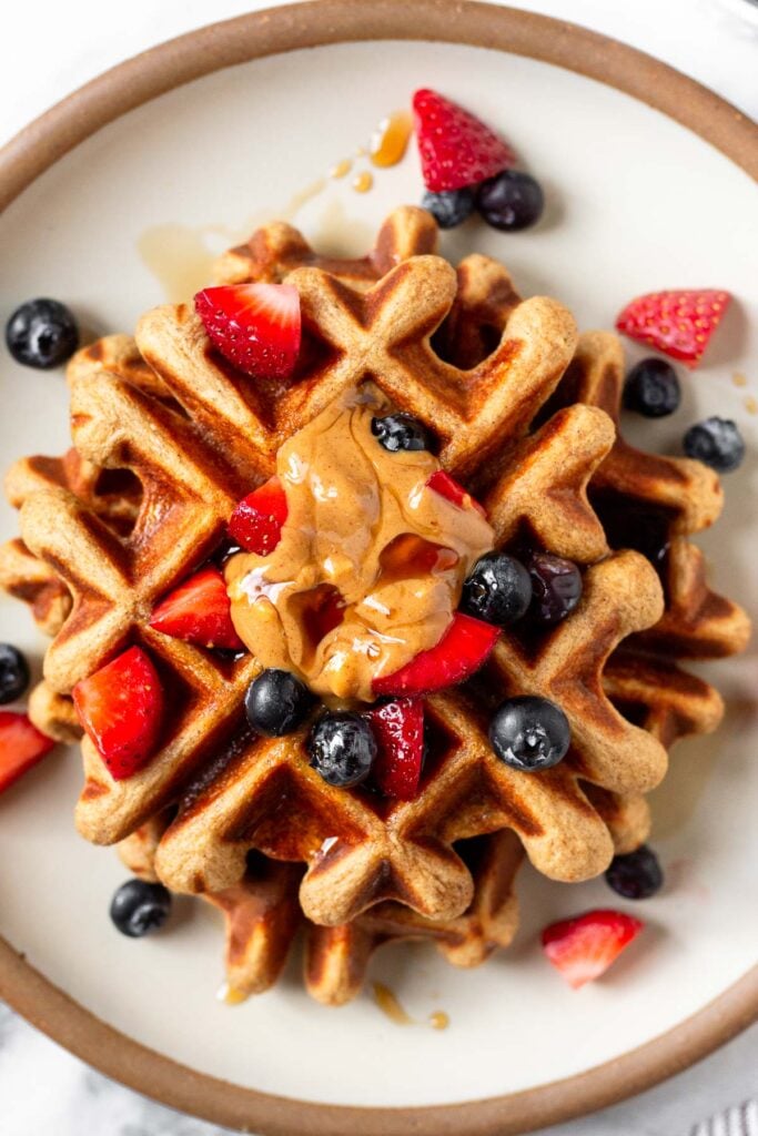 Overhead shot of protein powder waffles topped with berries, peanut butter, and maple syrup on a white plate.