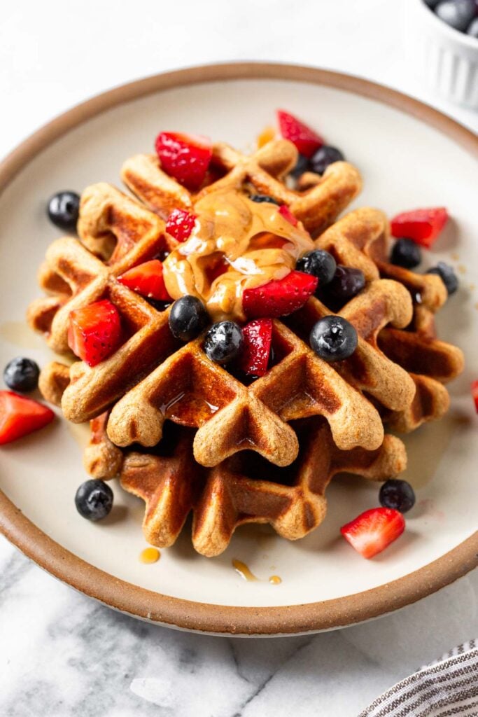 Two high protein waffles stacked on top of each other on a white plate. They are topped with strawberries, blueberries, peanut butter, and maple syrup.
