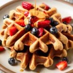 Protein waffles on a plate topped with berries and nut butter. Maple syrup is doing poured on top of them. Behind the plate is a small dish of berries.