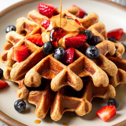 Protein waffles on a plate topped with berries and nut butter. Maple syrup is doing poured on top of them. Behind the plate is a small dish of berries.