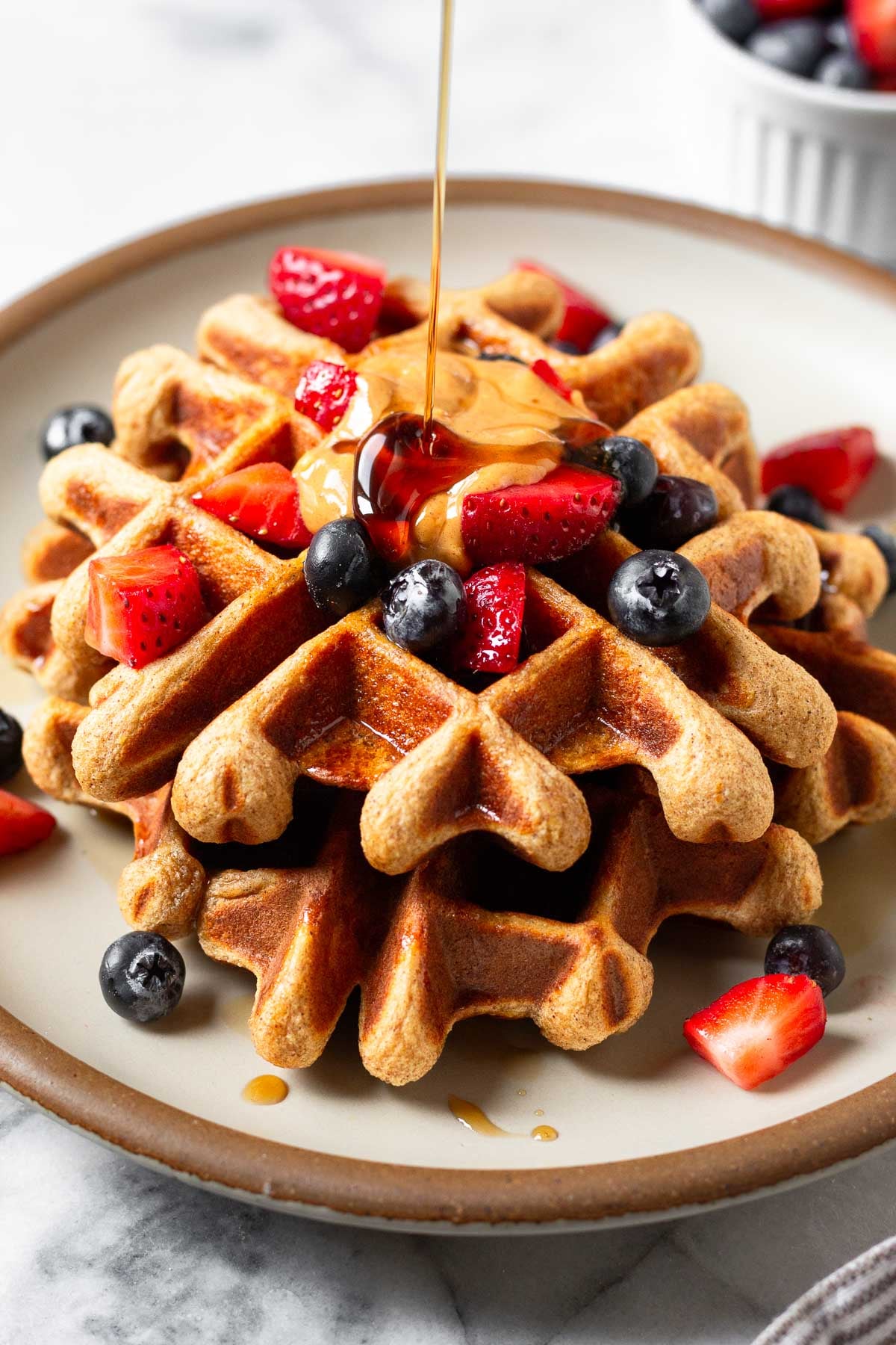 Protein waffles on a plate topped with berries and nut butter. Maple syrup is doing poured on top of them. Behind the plate is a small dish of berries.