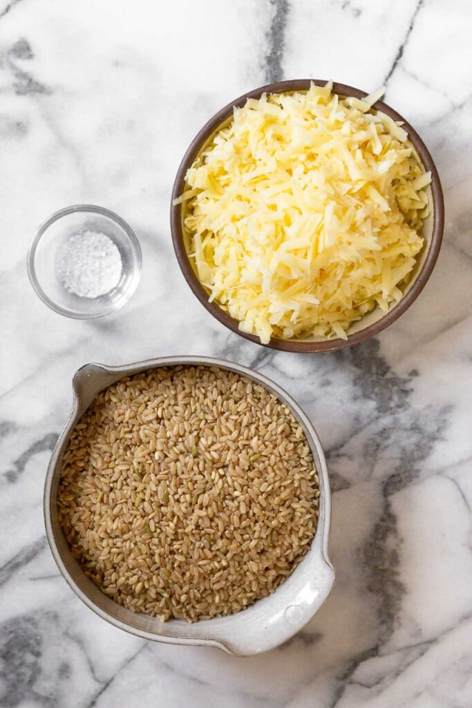 White marble counter with a bowl of shredded cheese, a bowl of brown rice, and a small bowl of salt.