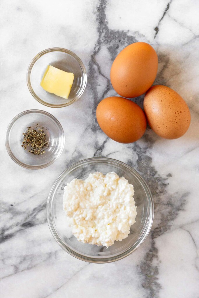 White marble counter with three brown eggs, a bowl of cottage cheese, a small bowl of pepper, and a bowl with a slice of butter in it.