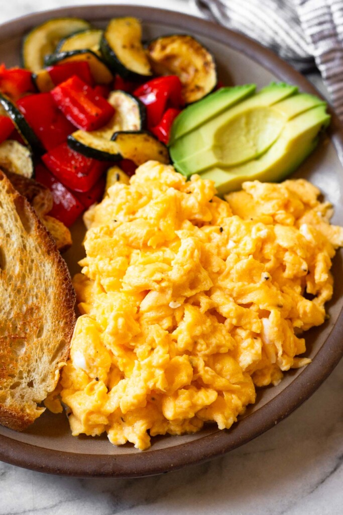 A close of up scrambled eggs with cottage cheese on a plate. Also on the plate is some toast, sautéed veggies, and avocado.
