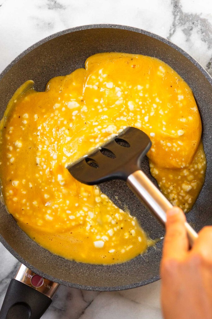 Large skillet with uncooked scrambled eggs in it. A spatula is pushing them in the pan.