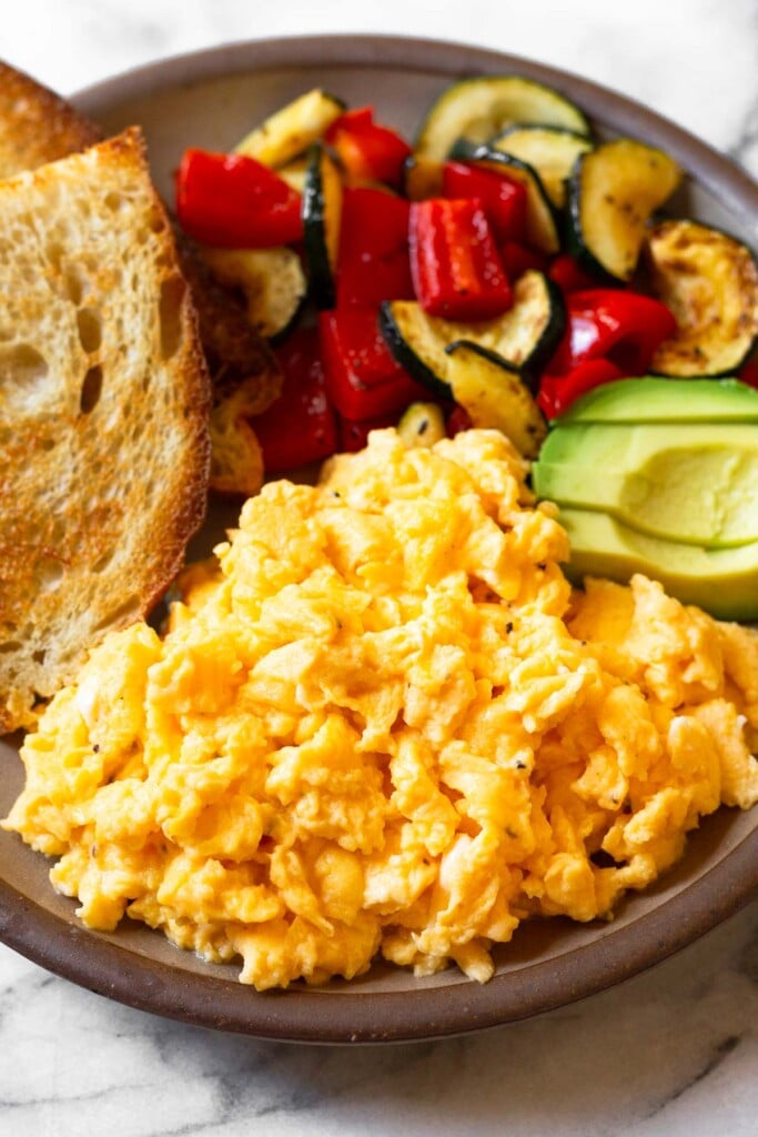 A close up a a plate with cottage cheese scrambled eggs, toast, sautéed veggies, and avocado.