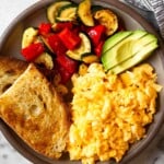 An overhead shot of a plate with scrambled cottage cheese eggs, two pieces of toast, sautéed veggies, and avocado slices.