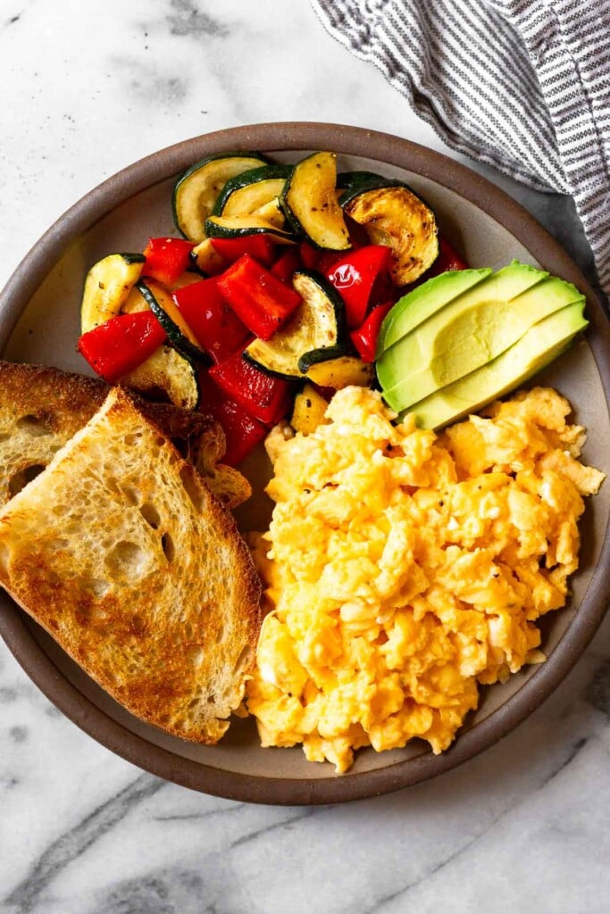 An overhead shot of a plate with scrambled cottage cheese eggs, two pieces of toast, sautéed veggies, and avocado slices.