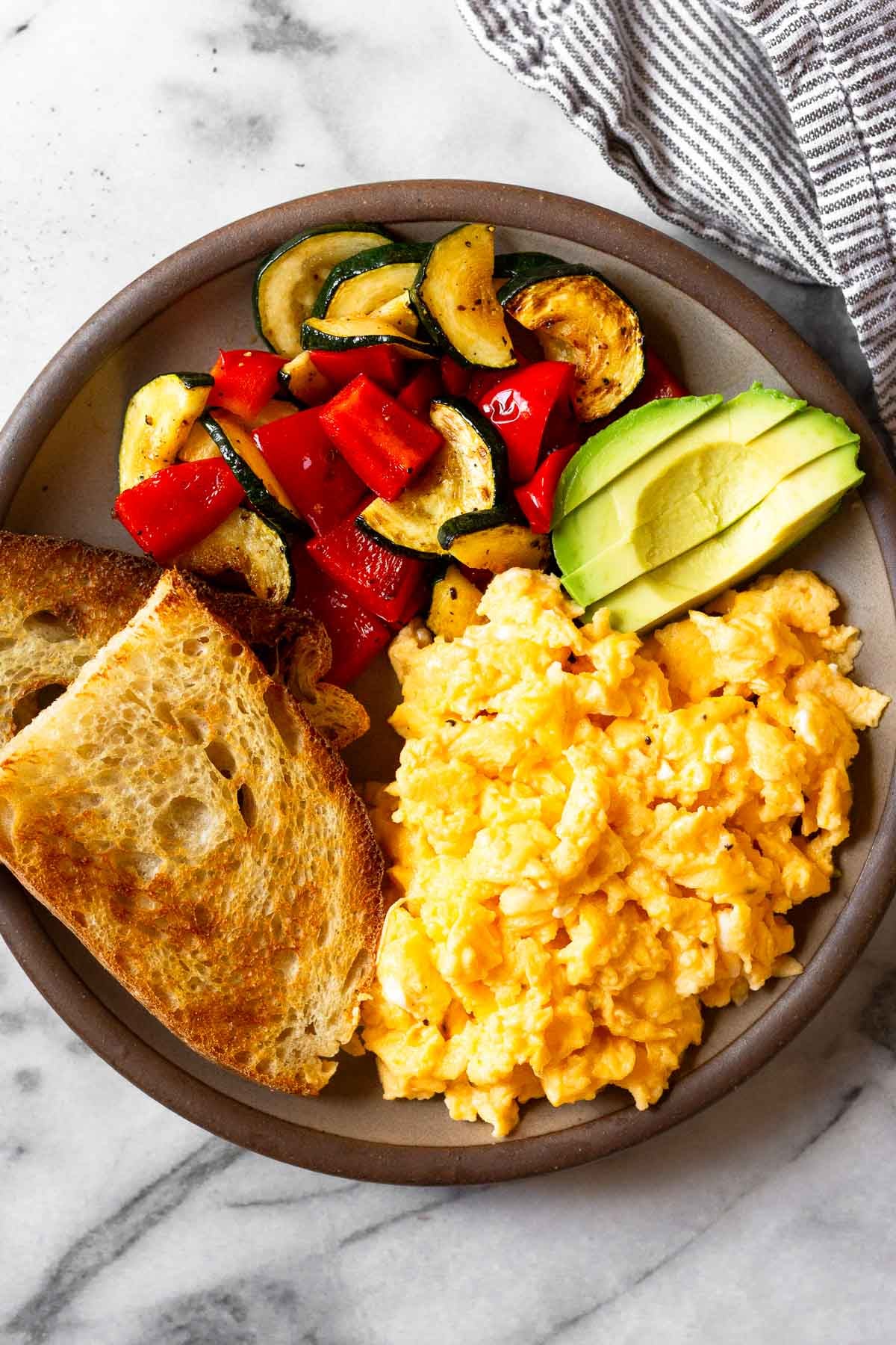 An overhead shot of a plate with scrambled cottage cheese eggs, two pieces of toast, sautéed veggies, and avocado slices.