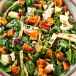 Chopped harvest salad with chicken, mixed greens, roasted sweet potatoes, dried cranberries, pumpkin seeds, and goat cheese all tossed with dressing in a white bowl. Next to the bowl is a jar of more dressing and a white kitchen towel.