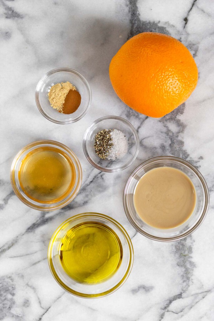 Mable counter top with a large orange, a bowl of salt and pepper, a bowl of tahini, a bowl of olive oil, a bowl of apple cider vinegar, and a bowl of spices.