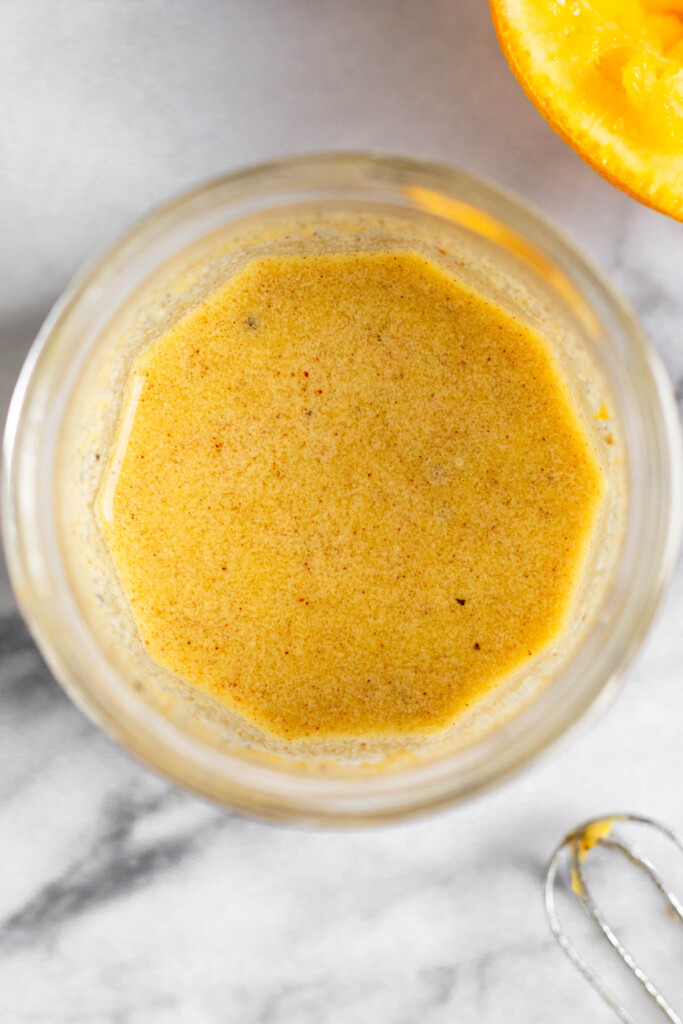 Overhead shot of a jar filled with an orange dressing. Next to it is a small whisk and a juiced orange.