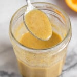 Orange vinaigrette in a jar with a spoon coming out of it. In front of the jar is a small whisk and behind the jar is half an orange.