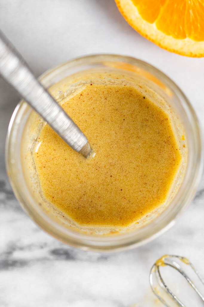 Overhead shot of orange juice vinaigrette in a jar with a spoon in it. Next to the jar is a small whisk and half of a cut open orange.