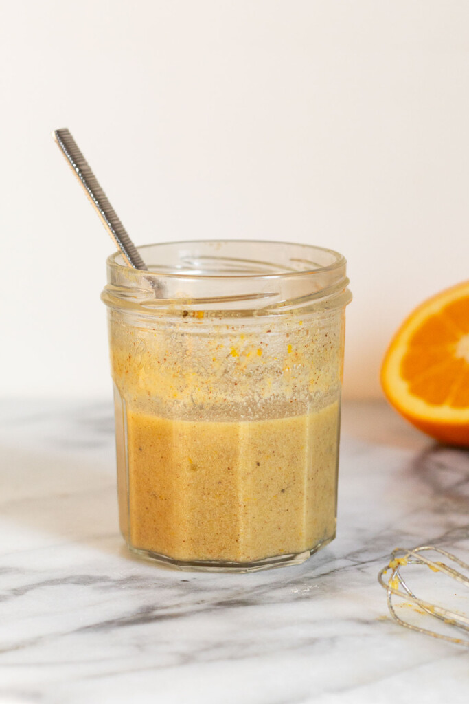 Orange vinaigrette dressing in a jar with a spoon in it. In front of the jar is a small whisk and behind the jar is half an orange.