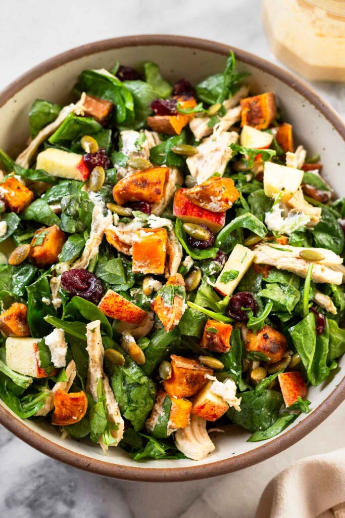 Large salad in a bowl with mixed greens, chicken, sweet potatoes, apples, cranberries, pumpkin seeds, and goat cheese all mixed together. Behind the bowl is a jar of dressing.
