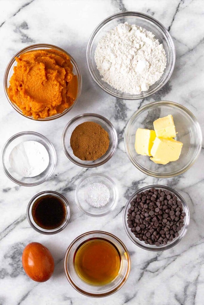 White marble counter with a bowl of flour, a bowl of butter, a bowl of chocolate chips, a bowl of maple syrup, one brown egg, a bowl of vanilla extract, a bowl of salt, a bowl of baking powder, a bowl of spices, and bowl of pumpkin puree.