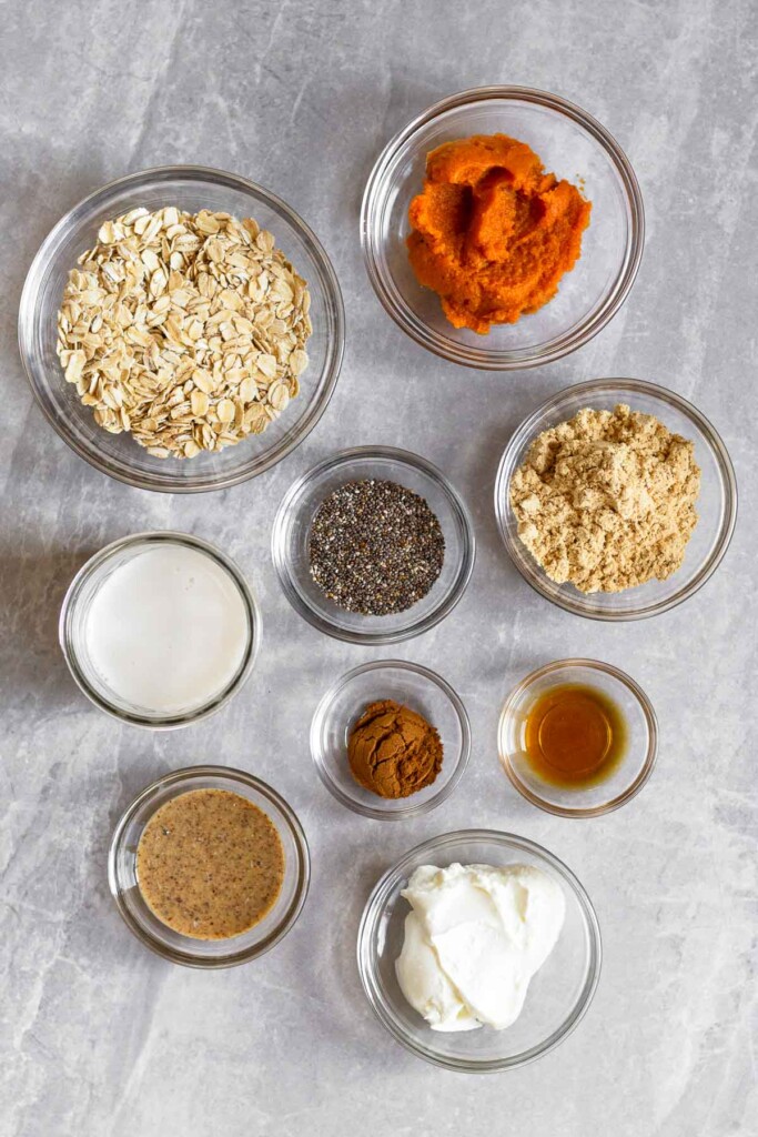 Grey marble counter top with a bowl of pumpkin puree, a bowl of protein powder, a bowl of vanilla extract, a bowl of yogurt, a bowl of almond butter, a bowl of spices, a bowl of chia seeds, and a bowl of rolled oats.
