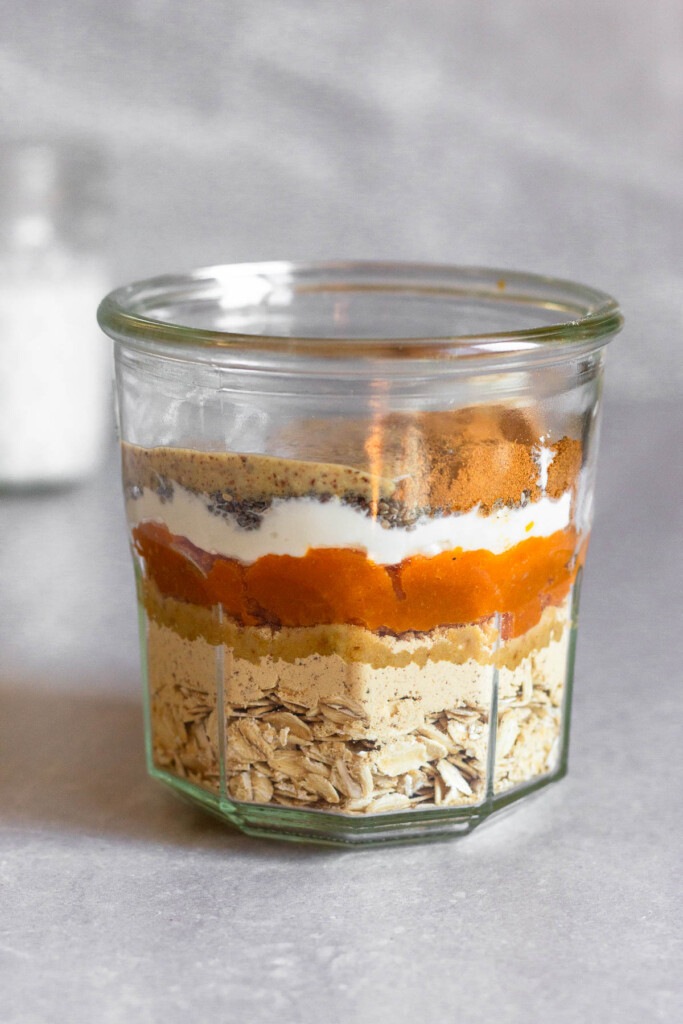 A straight on shot of a jar layered with rolled oats, protein powder, pumpkin puree, greek yogurt, chia seeds, almond butter, and spices. Behind it is a jar of milk.