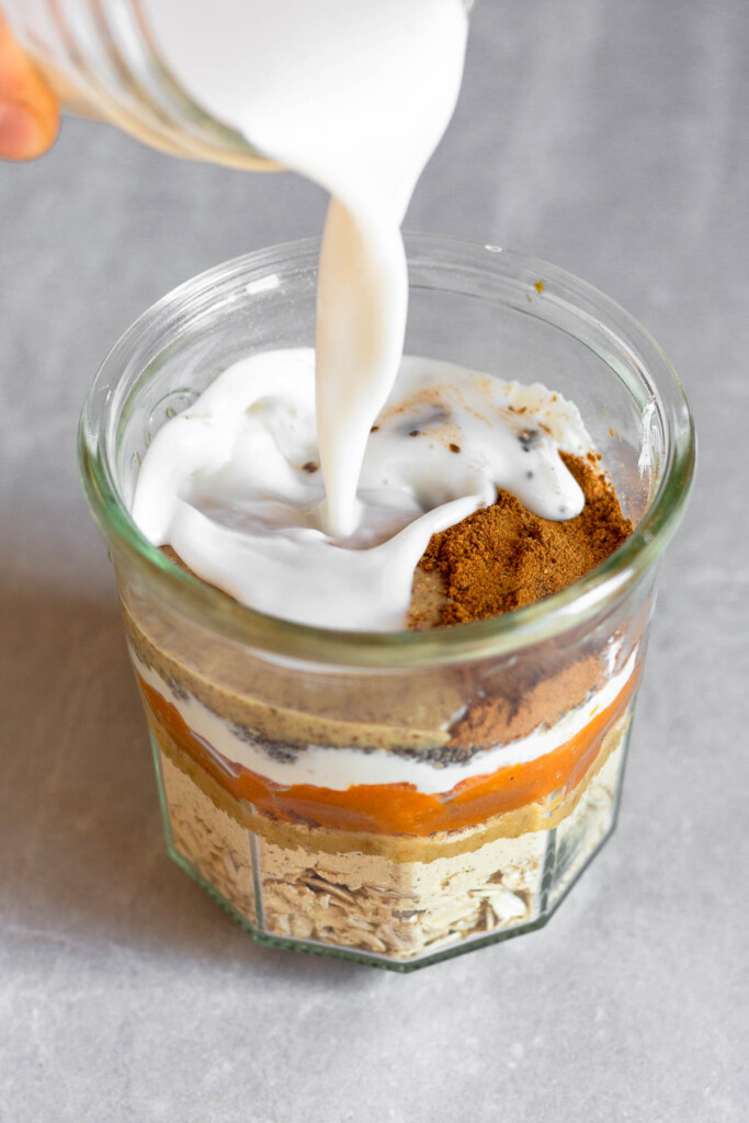 A jar layered with rolled oats, protein powder, pumpkin puree, greek yogurt, chia seeds, almond butter, and spices with milk being poured on top.