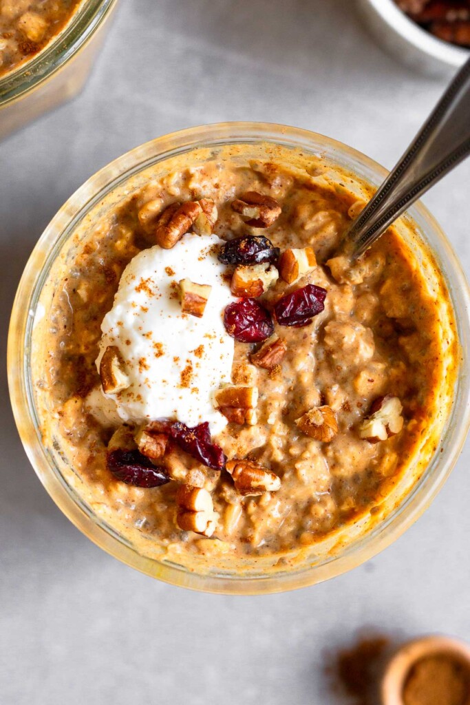 Overhead shot of a jar of pumpkin spice overnight oats topped with diced greek yogurt, chopped pecans, dried cranberries, and cinnamon with a spoon in the jar. Next to it is other jar of overnight oats, a teaspoon of cinnamon, and a ramekin of pecans.