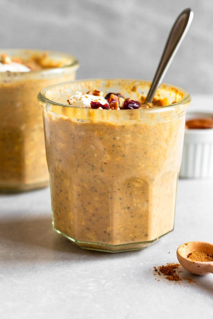 Two jars of pumpkin pie overnight oats, with one being behind the other. They are topped with toppings and a spoon is in the jar in the front. In front of the is a teaspoon of cinnamon and off to the back is a dish of pecans.