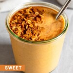 Sweet potato smoothie in a large jar with a metal straw in it topped with granola and cinnamon. In front of the jar is a teaspoon filled with cinnamon. Behind the jar is a bowl of granola and a white kitchen towel.