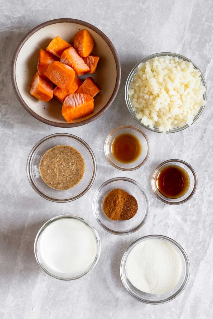 Grey marble counter with a bowl of frozen sweet potato cubes, a bowl of frozen cauliflower rice, a small bowl of vanilla extract, a small bowl of maple syrup, a bowl of spices, a bowl of protein powder, a jar of milk, and a bowl of almond butter.