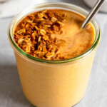 Sweet potato smoothie in a large jar with a metal straw in it topped with granola and cinnamon. In front of the jar is a teaspoon filled with cinnamon. Behind the jar is a bowl of granola and a white kitchen towel.