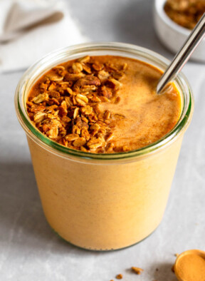 Sweet potato smoothie in a large jar with a metal straw in it topped with granola and cinnamon. In front of the jar is a teaspoon filled with cinnamon. Behind the jar is a bowl of granola and a white kitchen towel.