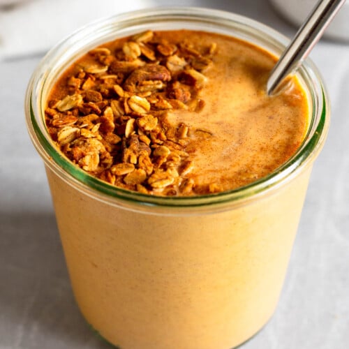 Sweet potato smoothie in a large jar with a metal straw in it topped with granola and cinnamon. In front of the jar is a teaspoon filled with cinnamon. Behind the jar is a bowl of granola and a white kitchen towel.