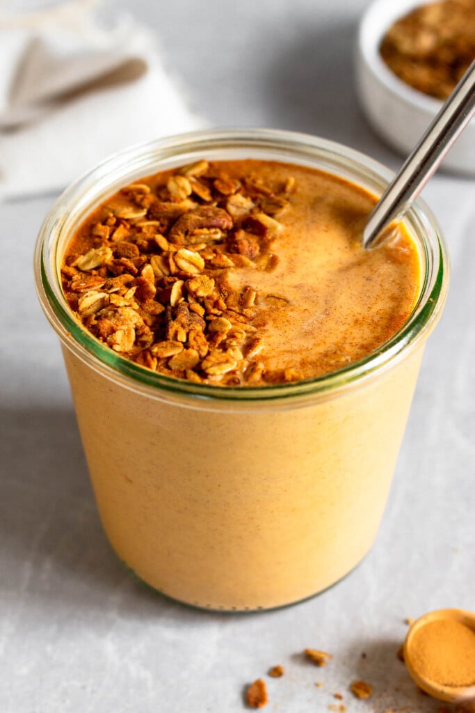 Sweet potato smoothie in a large jar with a metal straw in it topped with granola and cinnamon. In front of the jar is a teaspoon filled with cinnamon. Behind the jar is a bowl of granola and a white kitchen towel.