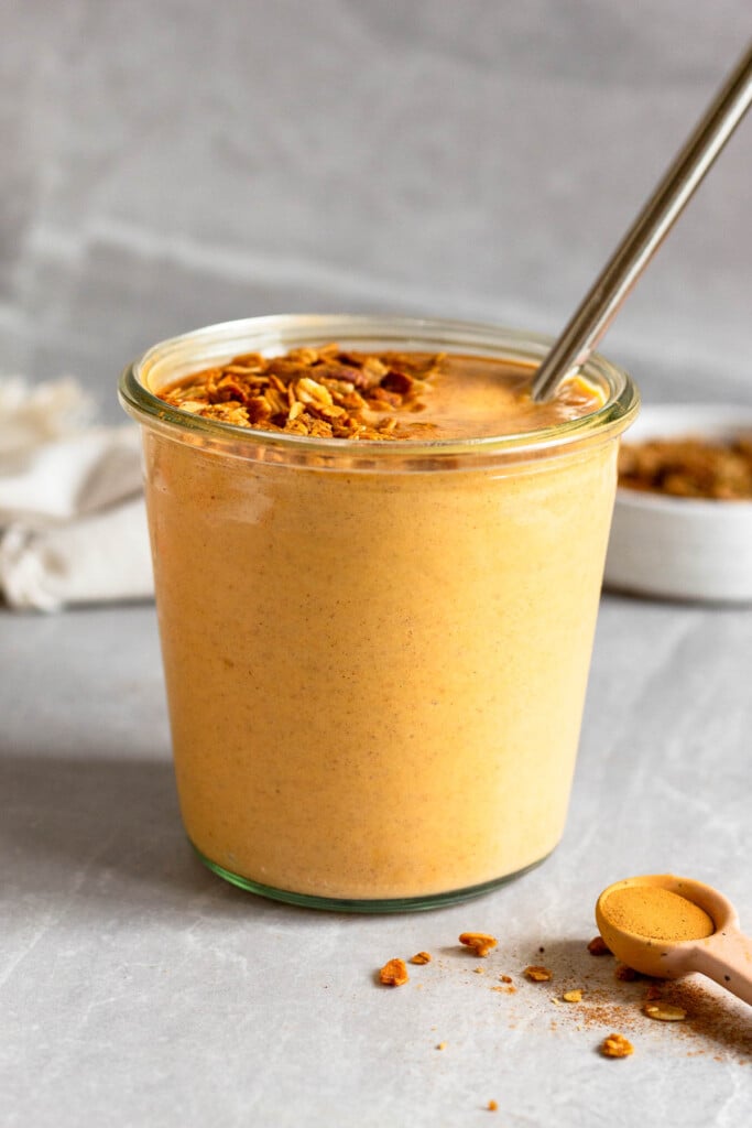 Sweet potato smoothie in a jar topped with granola and a metal straw in it. In front of it is teaspoon filled with cinnamon and behind it is a bowl of granola and a white towel.