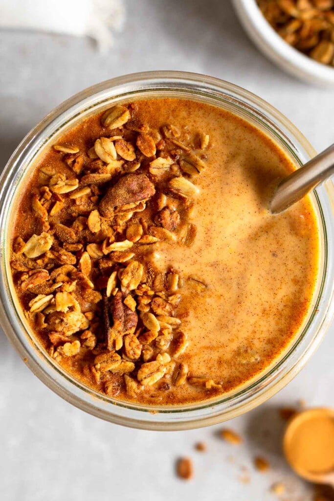 Overhead shot of a sweet potato smoothie topped with granola and cinnamon with a spoon stuck in it. Around it is a teaspoon filled with cinnamon, a bowl of granola, and a white kitchen towel.