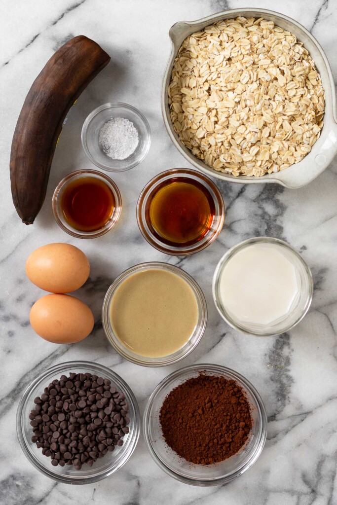 White marble counter with a bowl of rolled oats, a bowl of maple syrup, a jar of milk, a bowl of cocoa powder, a bowl of chocolate chips, a bowl of tahini, 2 brown eggs, a bowl of vanilla extract, a bowl of salt, and a brown banana.