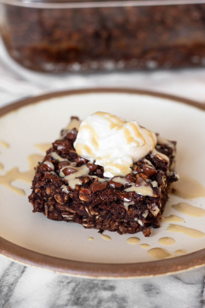 A piece of chocolate baked oatmeal on a plate topped with yogurt and nut butter. Behind it is the pan of the rest of the oatmeal.