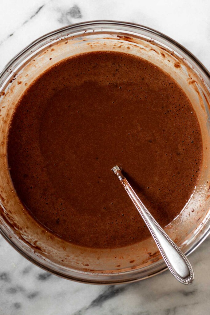 Wet chocolate batter in a medium glass bowl with a fork in it on a white marble counter.