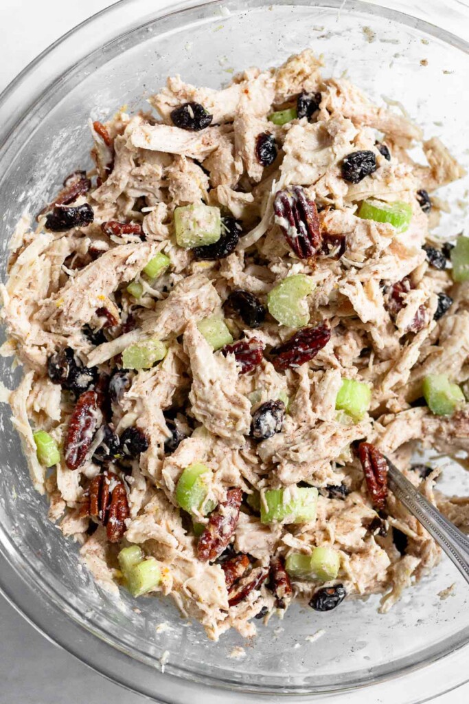 A large glass bowl of chicken salad with cranberries, pecans, and celery with a spoon in it.