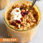 A jar of pumpkin overnight oats topped with greek yogurt, chopped pecans, dried cranberries, and cinnamon. A spoon is in the jar in the front. Around it is a ramekin of pecans, a teaspoon on cinnamon, and another jar of overnight oats.