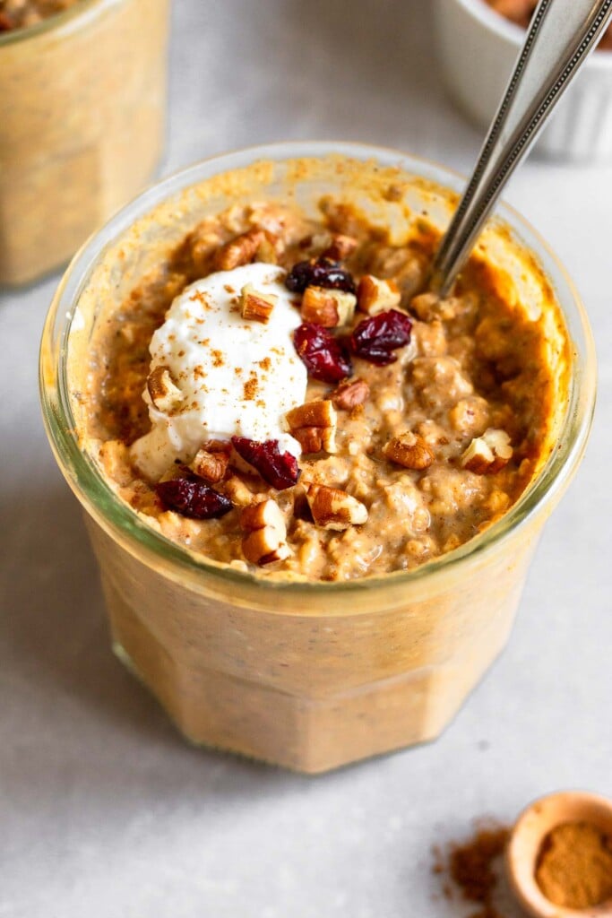 Healthy pumpkin overnight oats recipe in a jar topped with greek yogurt, chopped pecans, dried cranberries, and cinnamon. A spoon is in the jar in the front. Around it is a ramekin of pecans, a teaspoon on cinnamon, and another jar of overnight oats.