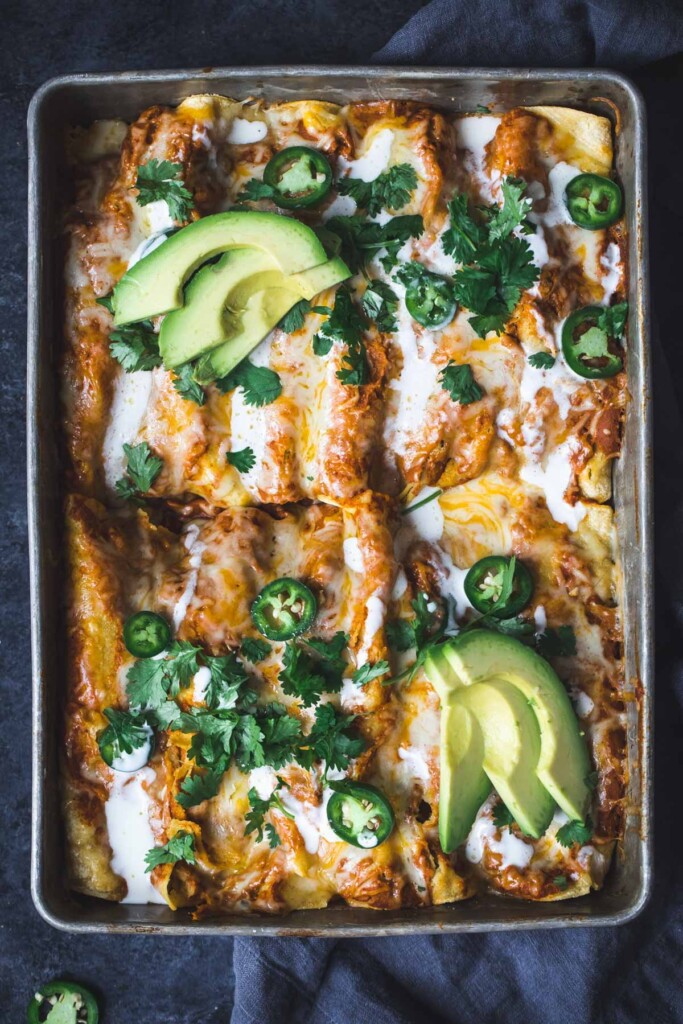 A dish of enchiladas topped with sour cream. sliced jalapeños, cilantro, and sliced avocado.